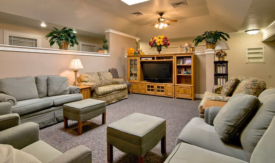 A cozy living room with multiple sofas and armchairs arranged around two ottomans. A wooden entertainment center with a TV, books, and decorative items is against the far wall. The room is warmly lit with a table lamp and ceiling fan lights, and decorated with plants and flowers.