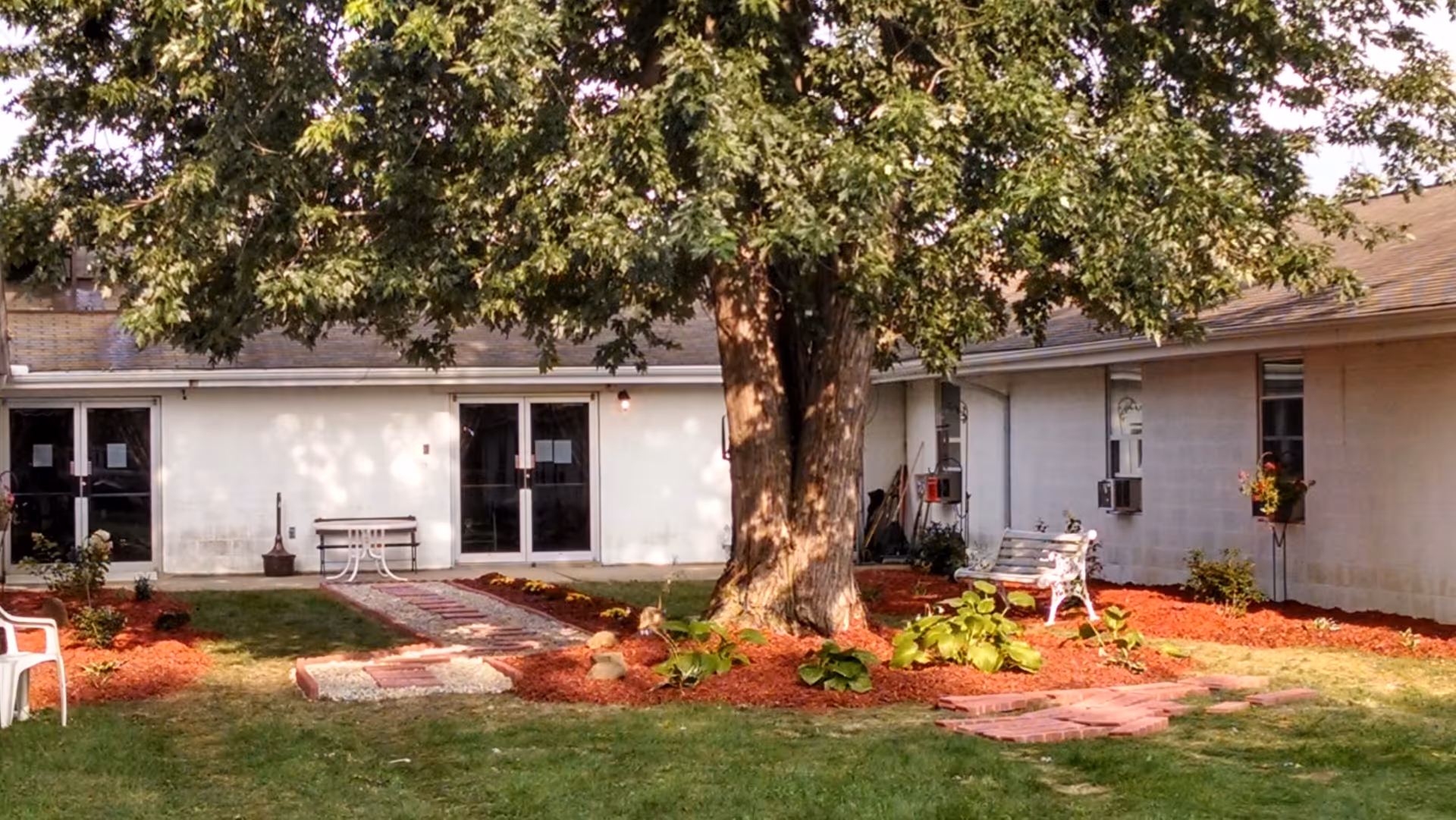 Outdoor courtyard area at Rock Falls Rehabilitation Center featuring a large tree in the center surrounded by landscaped flower beds with red mulch. There are white benches and plastic chairs placed around the courtyard, with a paved walkway leading to glass doors of the building.