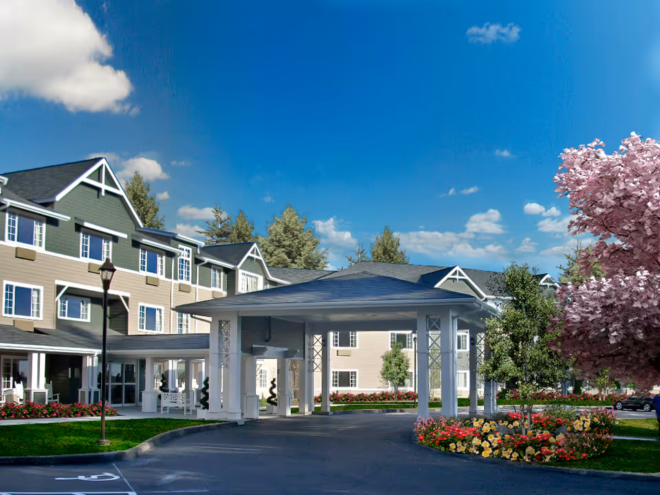 Exterior view of Ledgewood Bay Assisted Living facility showing a large covered entrance with white pillars, a multi-story building with green and beige siding, several windows, a lamppost, and landscaped grounds with colorful flowers and trees including a pink flowering tree under a bright blue sky with some clouds.