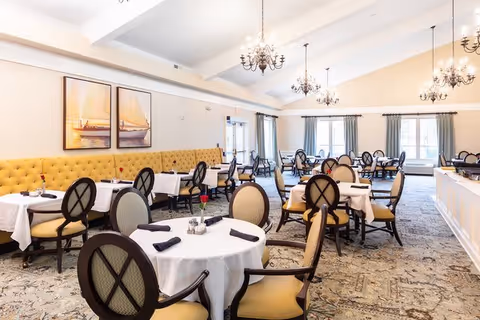 A bright and spacious dining room with multiple round tables covered with white tablecloths, each set with black napkins and a single red rose in a small vase. The room features elegant chandeliers hanging from a high ceiling, large windows with blue curtains, and a long yellow tufted bench along one wall. The carpet has a patterned design, and the overall atmosphere is inviting and well-lit.