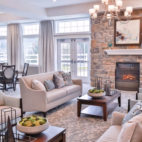 A bright and cozy living room with beige sofas and armchairs arranged around a dark wooden coffee table. The room features a stone fireplace with a lit fire, a modern chandelier, large windows with curtains, and a dining table with chairs in the background. Decorative plants and artwork add to the inviting atmosphere.