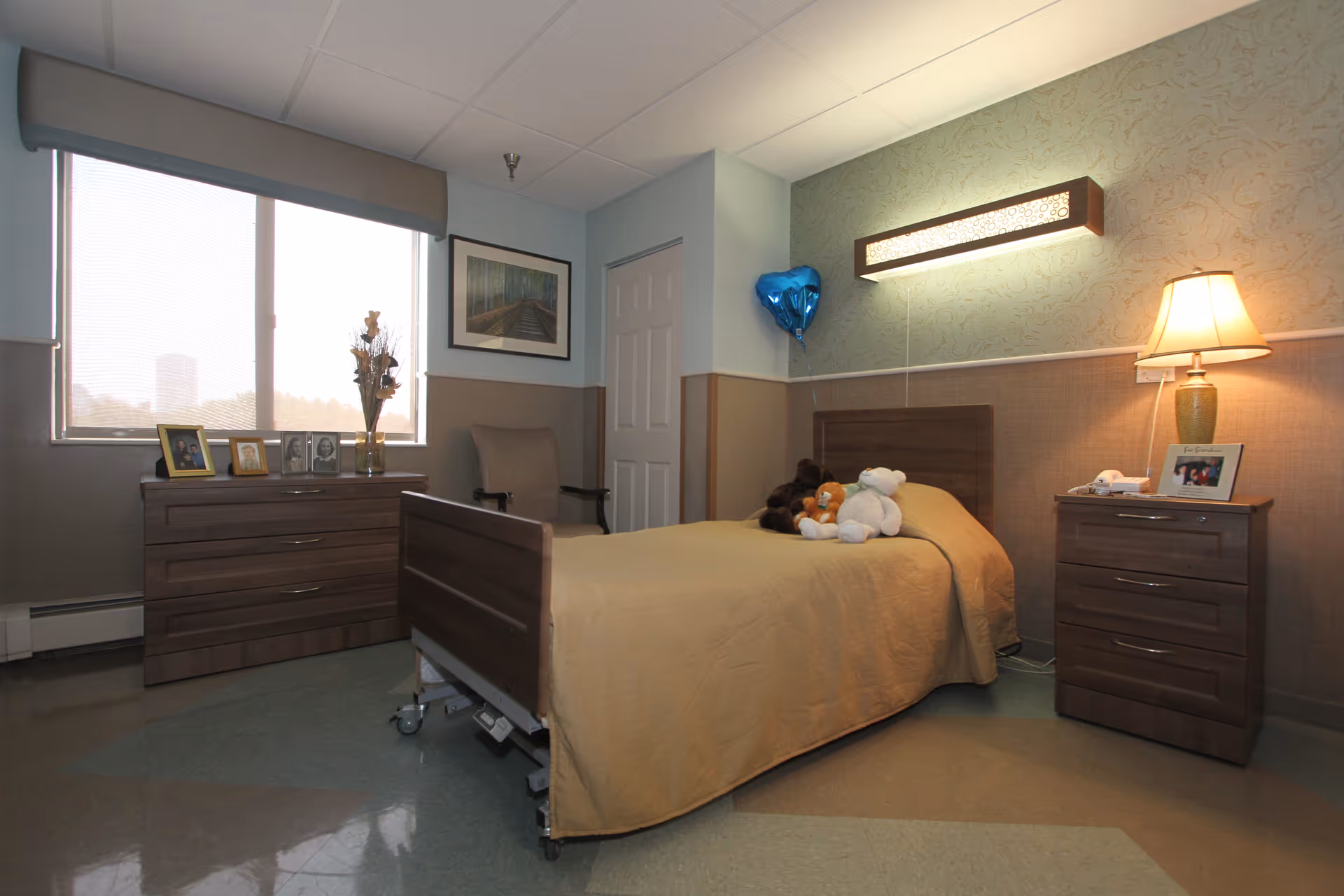 A small bedroom in a senior living facility with a single bed covered in a beige blanket and several stuffed animals on it. There is a wooden dresser with framed photos and a vase with dried flowers next to a large window with blinds. A cushioned chair is placed near a closed door. On the right side, there is a wooden nightstand with a lamp and a framed photo. The walls are painted in two tones with a decorative light fixture above the bed and a blue heart-shaped balloon attached to the wall.
