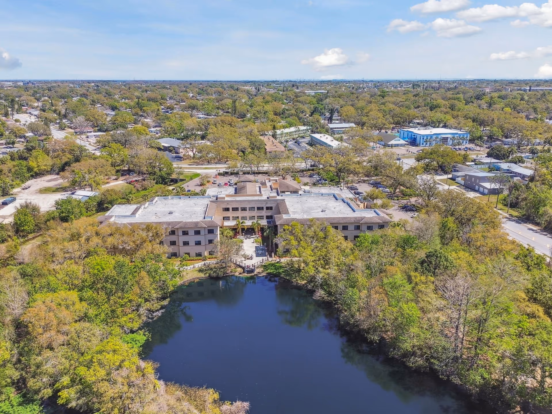 Aerial view of The Preserve at Dunedin facility showing a large building surrounded by trees and greenery with a pond in the foreground. The surrounding area includes roads, parking lots, and residential neighborhoods under a partly cloudy sky.