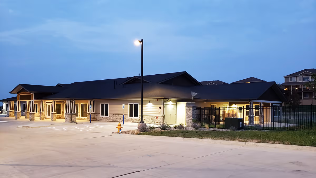 Exterior view of a single-story assisted living facility building at dusk with lights on, a parking lot with handicap spaces, a street lamp, and a fenced area on the right side.