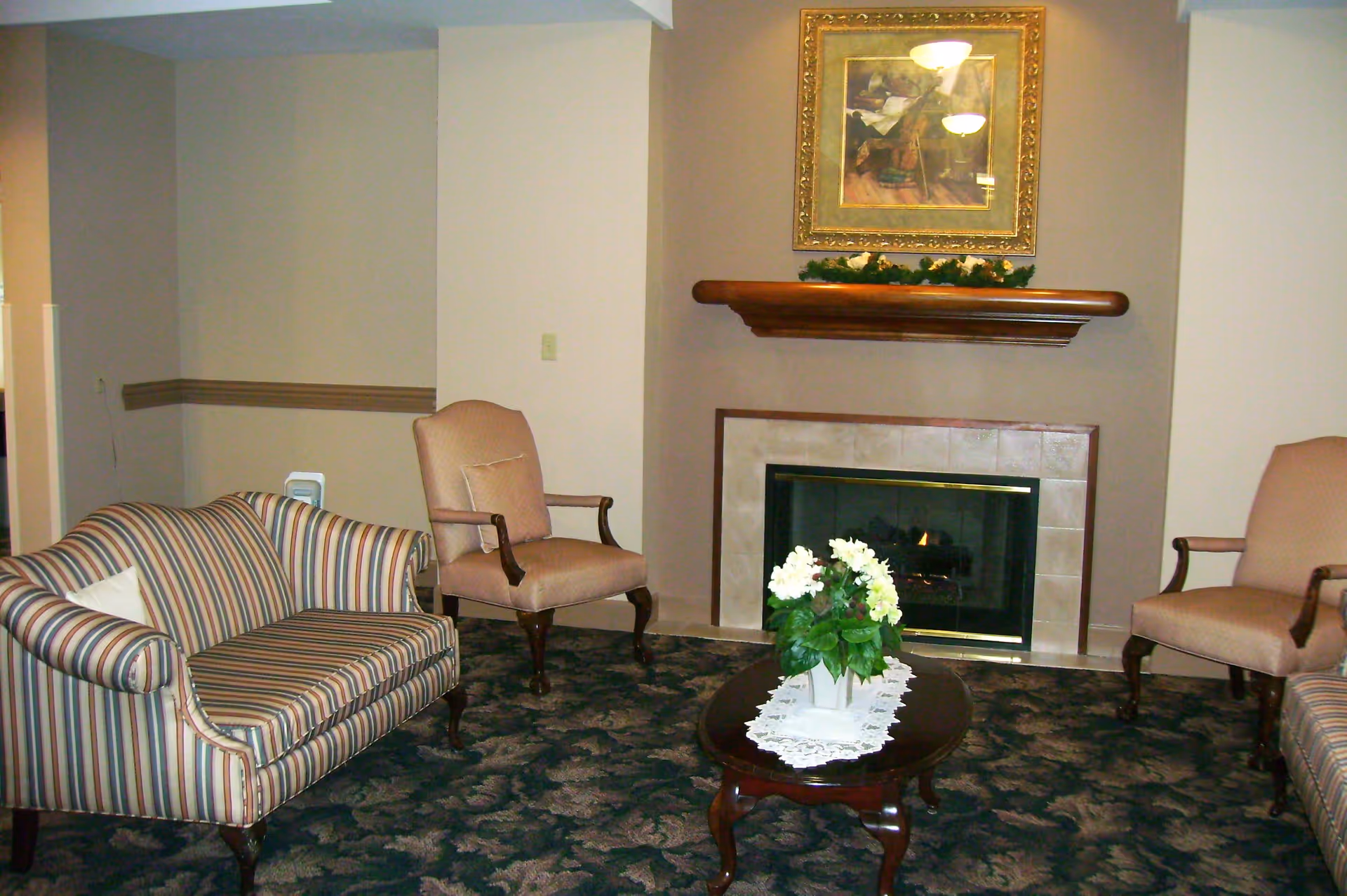 A cozy living room with a striped loveseat, two beige armchairs, a dark wooden coffee table with a white lace doily and a flower arrangement, and a fireplace with a wooden mantel and a framed painting above it.