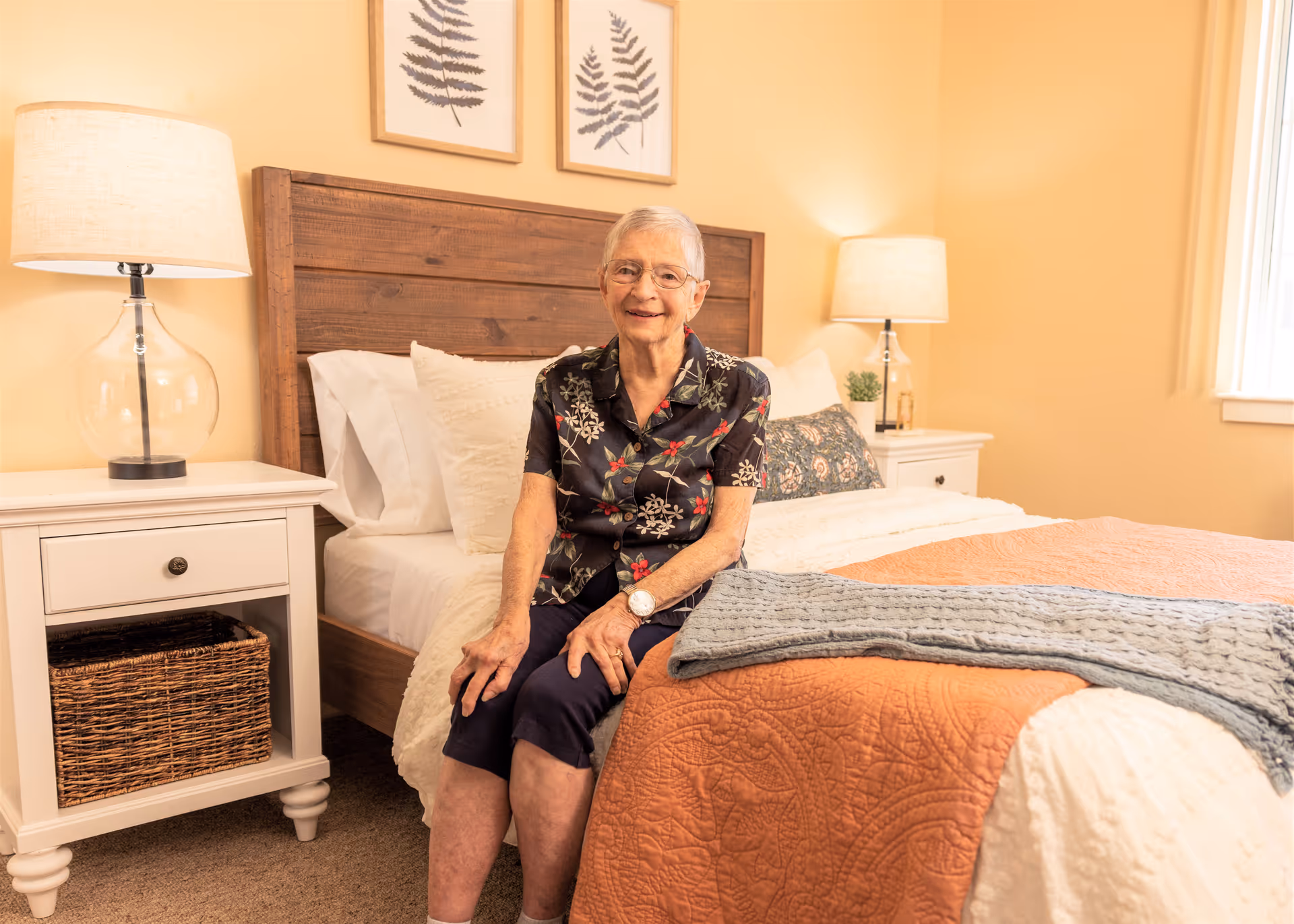 An elderly woman sits on the edge of a neatly made bed in a warmly lit bedroom with bedside tables and lamps.
