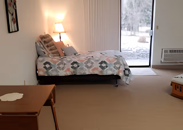 A bedroom with a single bed covered in a patterned quilt and several pillows. A lit table lamp is on a nightstand next to the bed. There is a sliding glass door leading outside, showing a snowy outdoor scene. A wooden table is partially visible in the foreground.