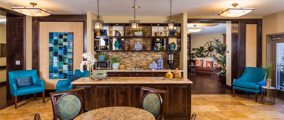 A cozy and well-decorated common area in a senior living facility featuring a kitchen island with decorative vases, a tiled backsplash, and open shelving with plants and pottery. The room includes turquoise armchairs, a round table with chairs, and a seating area with a brown leather sofa, plants, and a large wall clock. Warm lighting fixtures hang from the ceiling, and the floor is tiled.