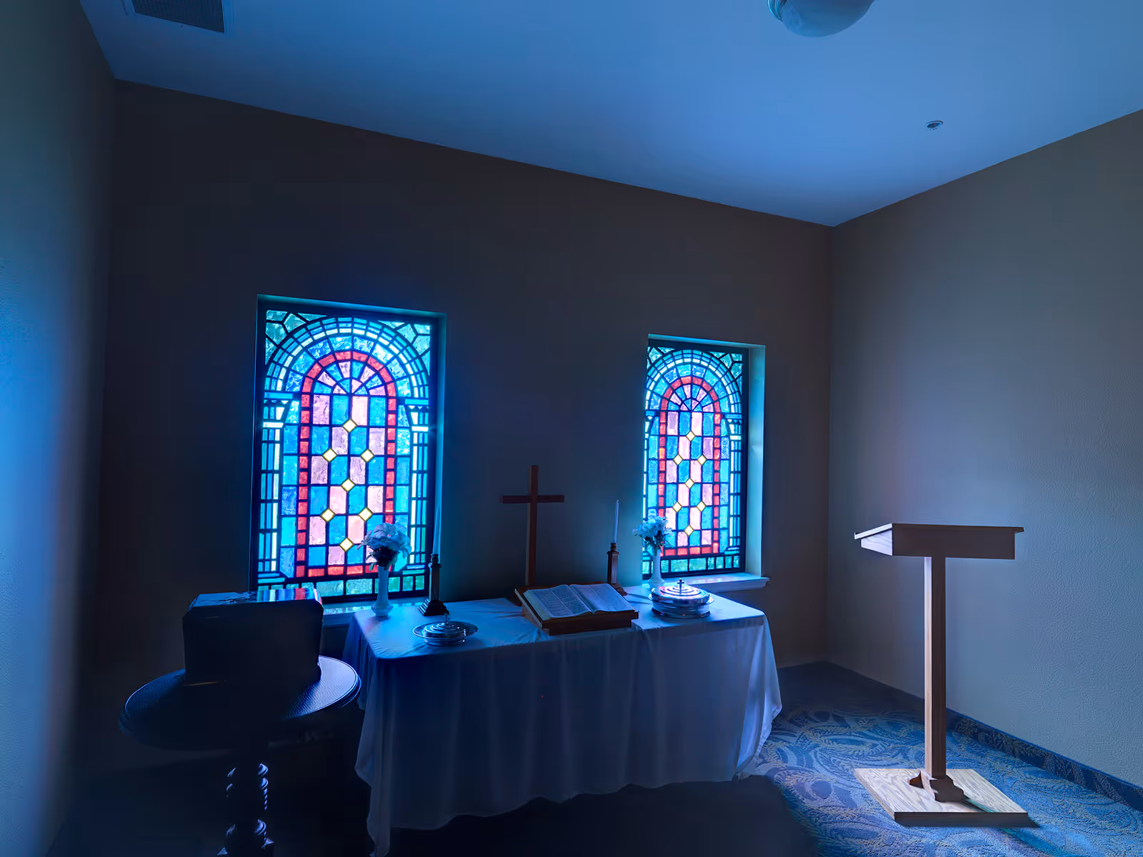 A small chapel room with two stained glass windows casting colorful light. There is a table covered with a white cloth holding a cross, an open Bible, candles, flowers, and communion plates. A wooden lectern stands to the right on a patterned carpeted floor.