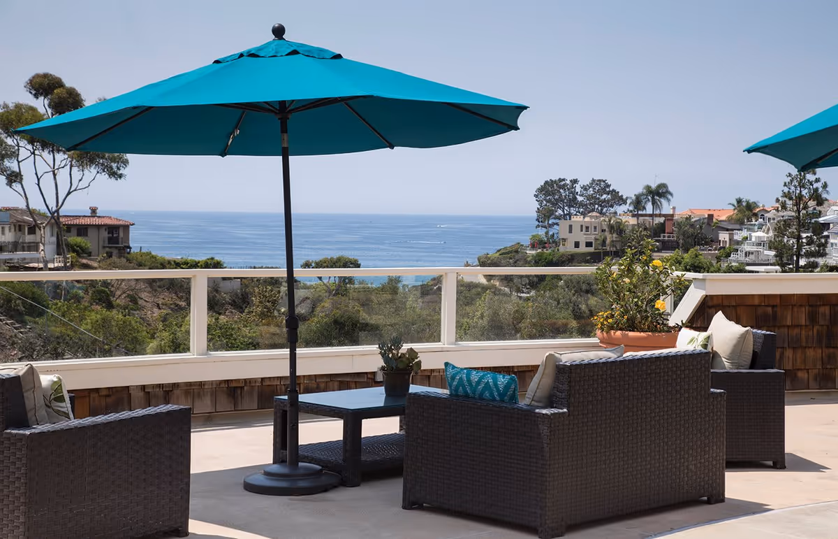 Outdoor patio area with dark wicker furniture including chairs and a coffee table, shaded by large teal umbrellas. The patio overlooks a scenic view of the ocean, trees, and nearby houses under a clear sky.