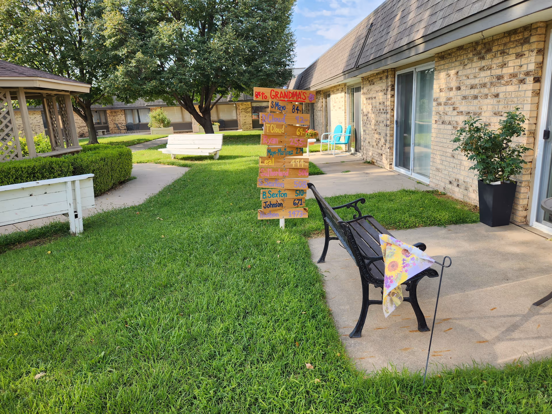 Outdoor courtyard area of a retirement village with green grass, trees, and benches. A wooden signpost with colorful handwritten names and numbers is placed in the center. There are brick buildings with sliding glass doors and patio chairs visible in the background.