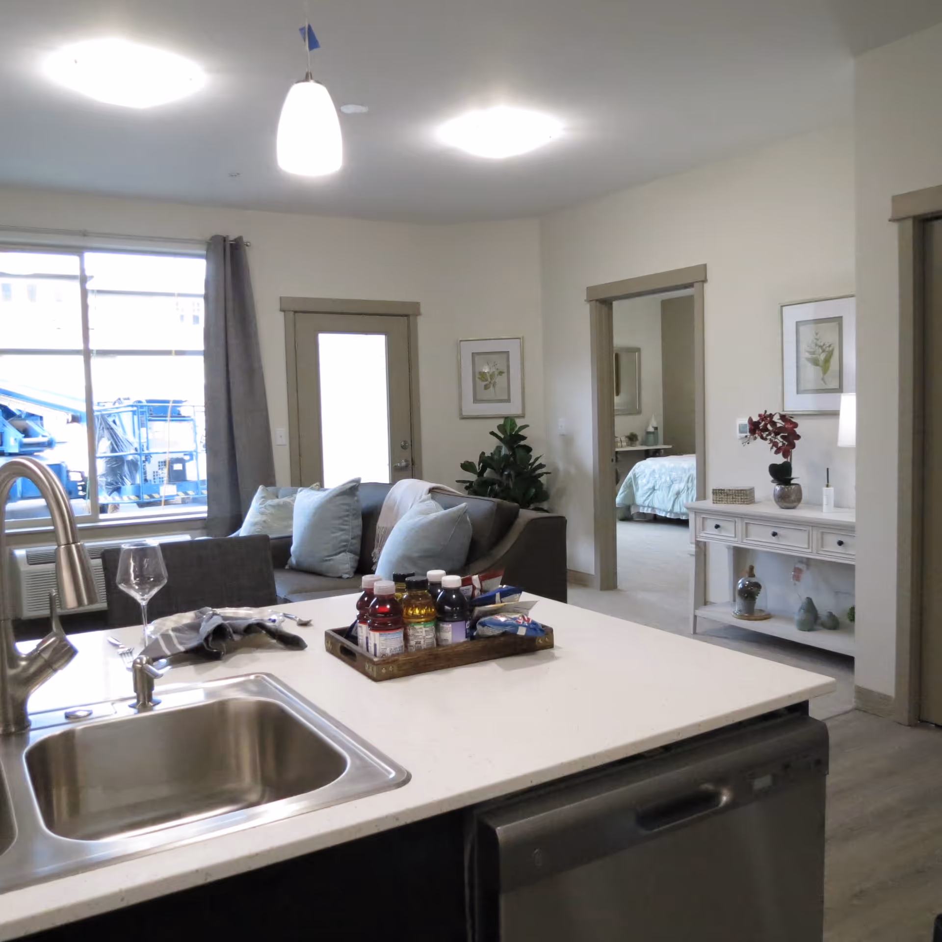 Open-plan apartment interior showing a kitchen island and sink in the foreground with a living area and a bedroom visible in the background.