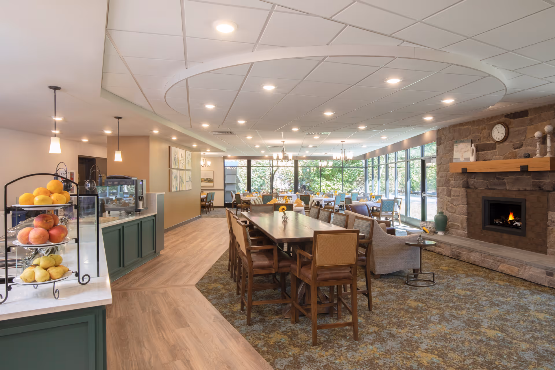 A spacious and well-lit dining area in a senior living facility with large windows allowing natural light to fill the room. The room features a long wooden dining table with chairs, a cozy seating area with armchairs near a stone fireplace with a clock above it, and several smaller dining tables in the background. There is a counter with a display of fresh fruit and a coffee machine on the left side. The ceiling has recessed lighting and a circular design element.