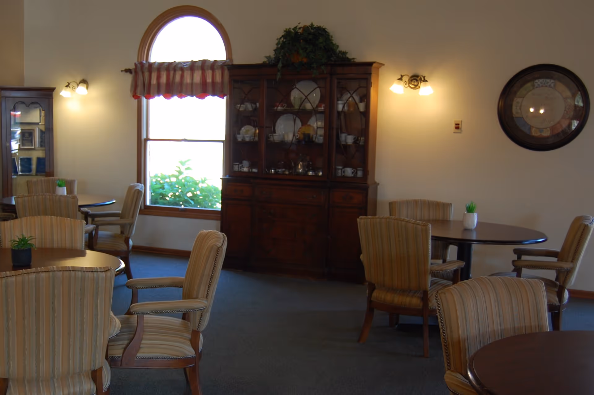 A cozy common room with round tables and striped upholstered chairs, a china cabinet, and a window.