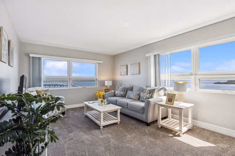A bright living room with large windows offering a view of the water and blue sky. The room features a gray sofa with patterned cushions, a white coffee table with a vase of yellow flowers, a side table with a lamp and framed photo, a patterned armchair, a TV, and a green plant. The walls are light gray and the floor is carpeted.