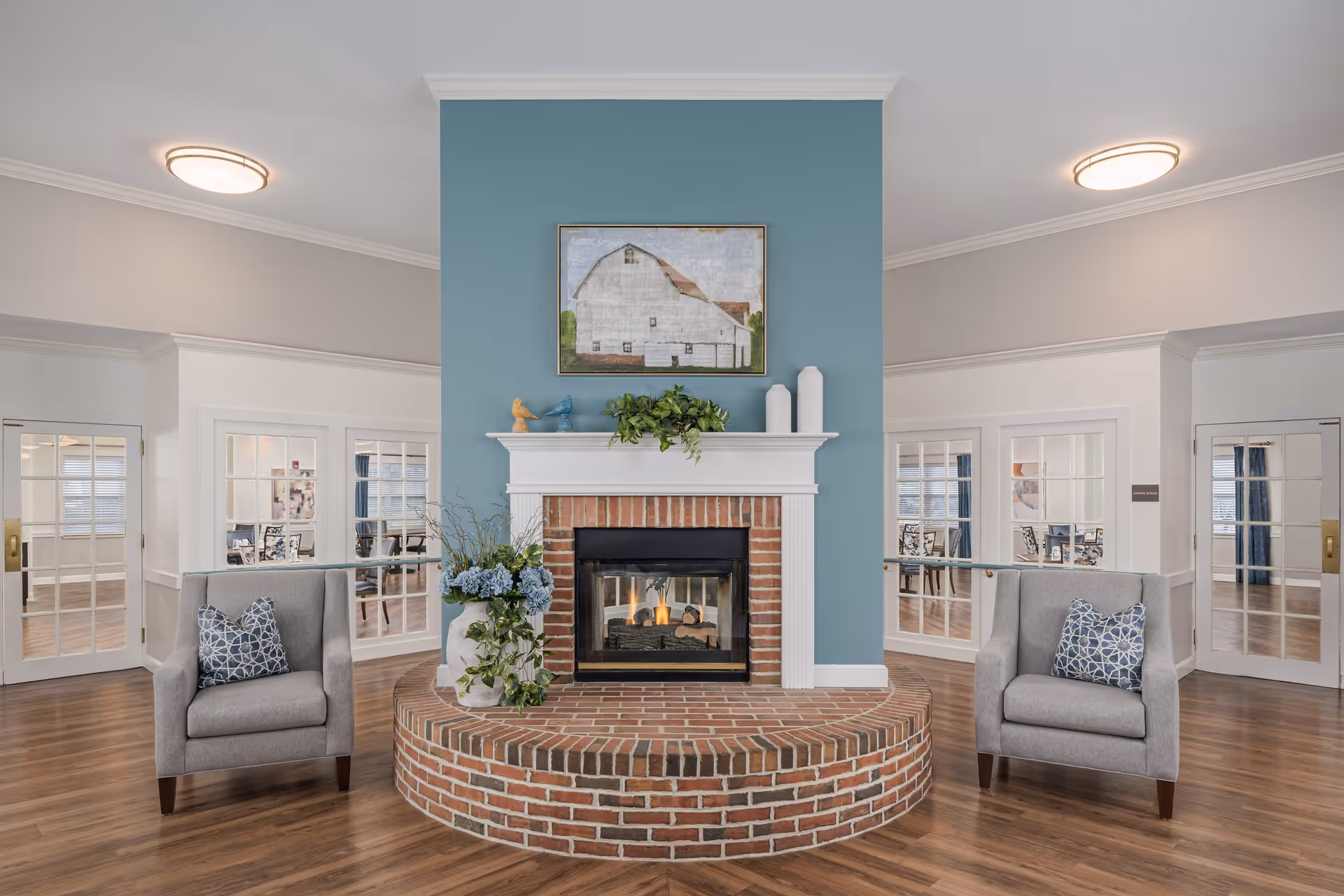 A cozy senior living common area featuring a central brick fireplace with a white mantel against a blue accent wall. Two gray armchairs with patterned blue pillows flank the fireplace. Above the mantel is a painting of a white barn. The room has wood flooring and white walls with crown molding. Behind the fireplace, glass-paneled double doors lead to a dining room.