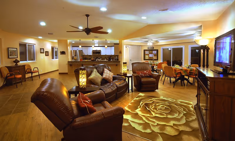 Warm, spacious common living area with leather sofas and armchairs, a TV, a dining table, and an open kitchen in the background.