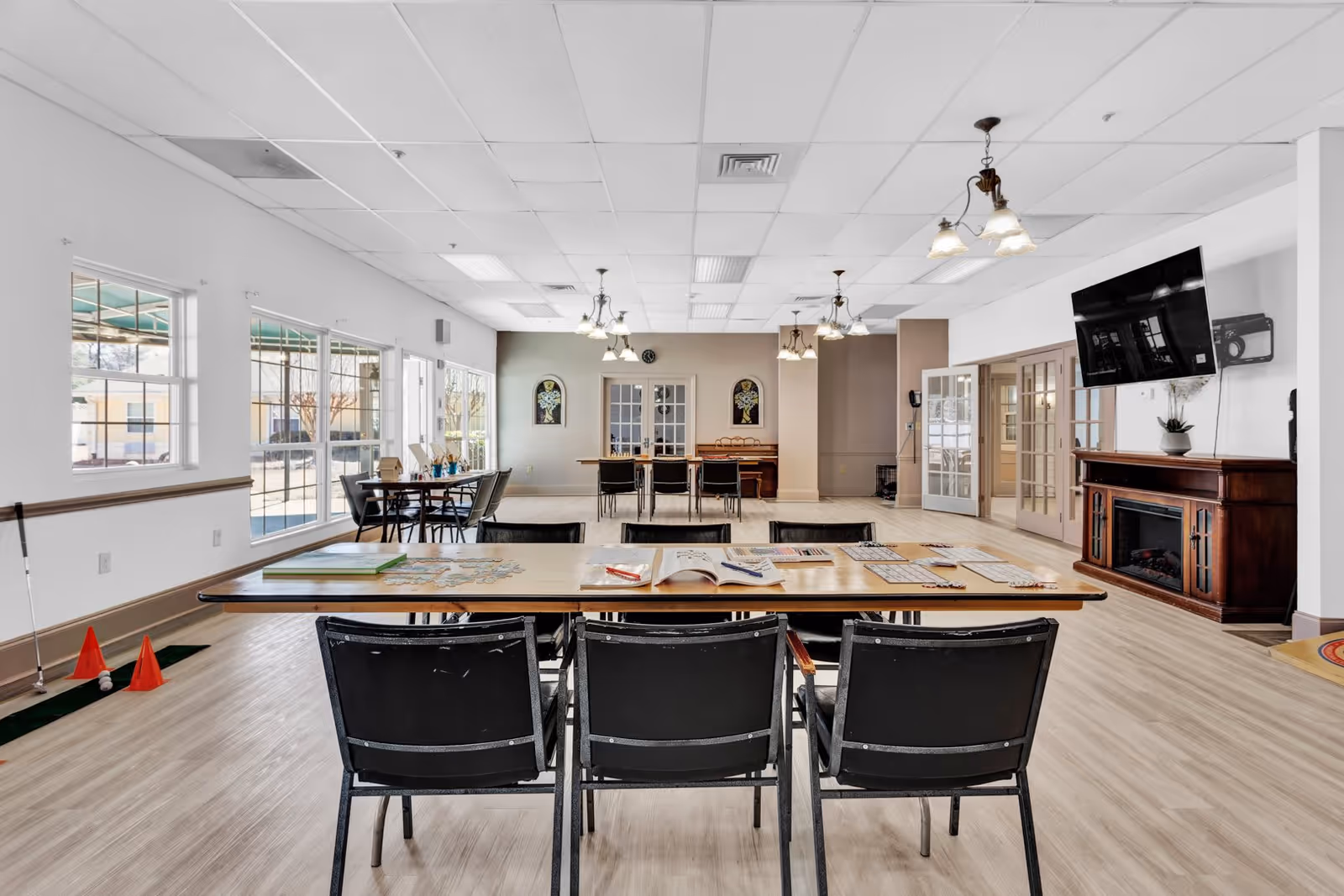 A spacious activity room in an assisted living facility with tables and chairs arranged for group activities. The tables have puzzles and books on them. Large windows on the left side let in natural light. There is a flat-screen TV mounted on the wall above a wooden cabinet with a fireplace insert. The room has light-colored flooring and ceiling lights with multiple bulbs.