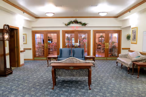 A spacious common area with blue patterned carpet, featuring two blue armchairs and a floral-patterned sofa arranged around a wooden coffee table. There is a grandfather clock on the left side and a floral arrangement above a set of glass double doors leading to another room with dining tables and chairs visible through the glass.