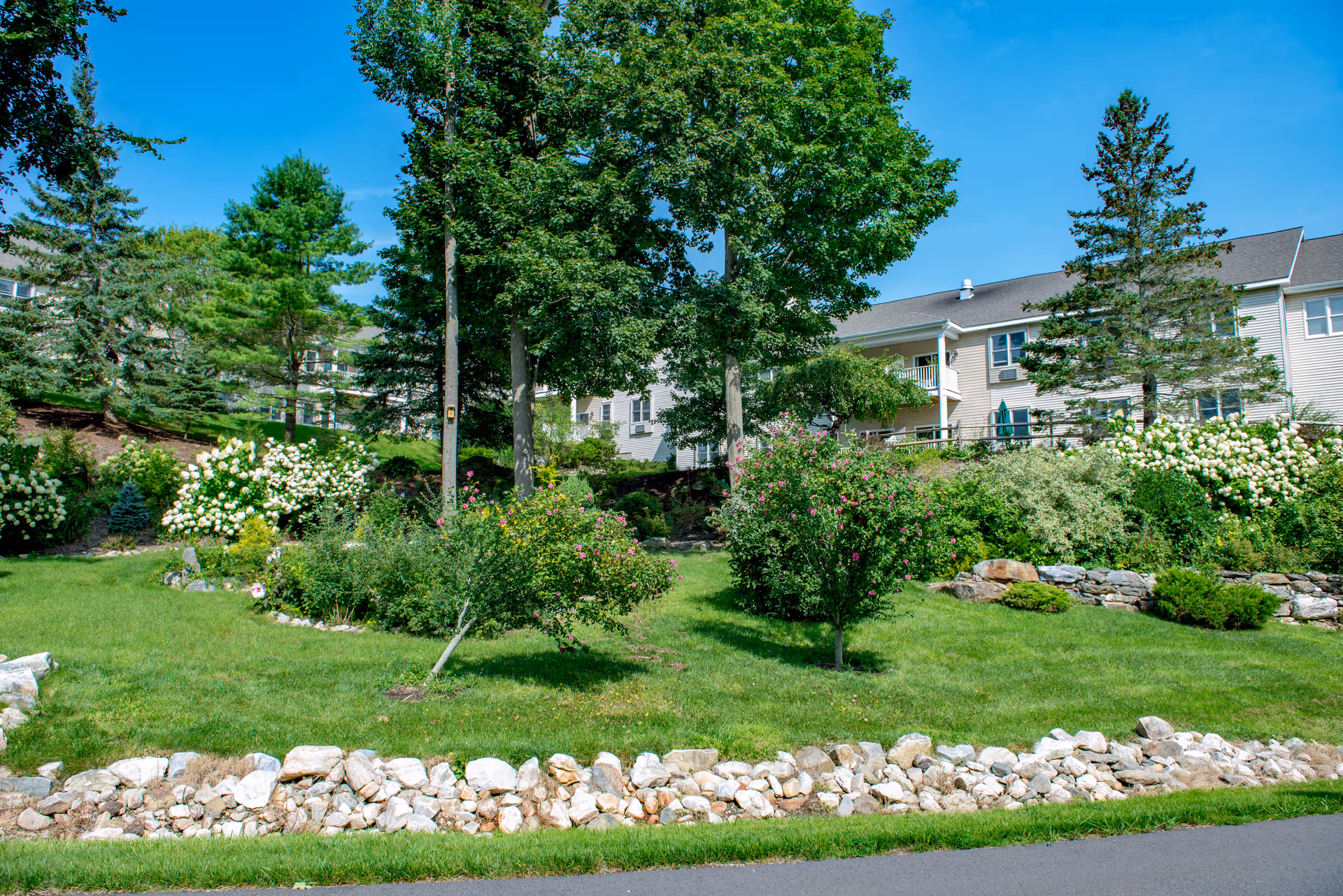 A landscaped outdoor area with green grass, various bushes, flowering plants, and several tall trees. In the background, there is a multi-story beige building with balconies and windows under a clear blue sky.