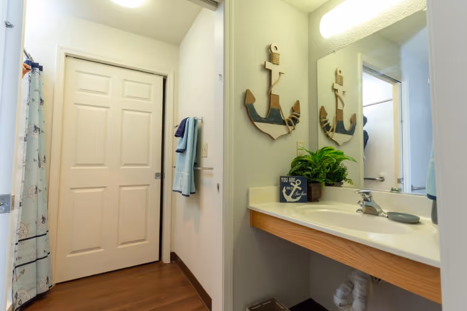 A bathroom with a white sliding door, a shower curtain with nautical designs, a towel rack with folded towels, a sink with a faucet, a large mirror, a decorative wooden anchor on the wall, a small potted plant, and a sign that reads 'You are my anchor'.
