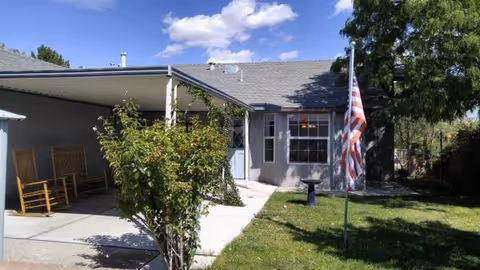 Single-story senior living facility exterior with a covered porch, chairs, and an American flag.