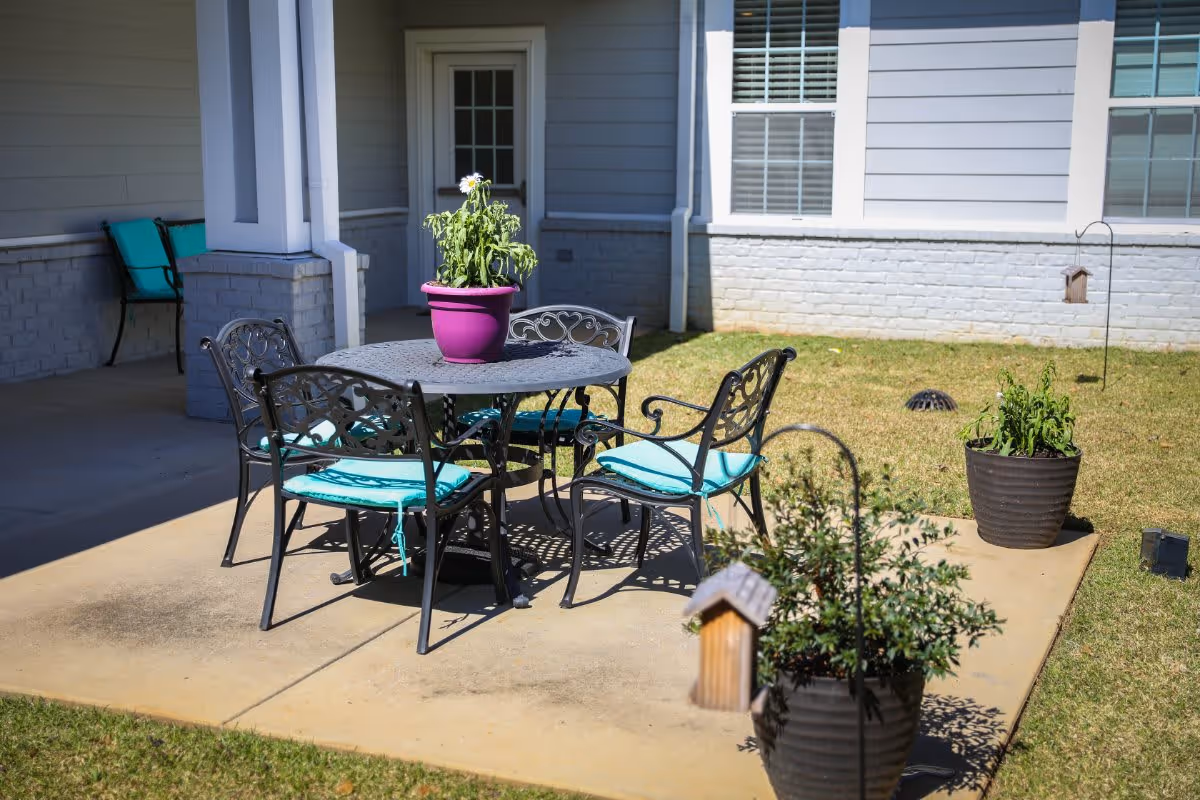 Outdoor patio area with a round metal table and four metal chairs with turquoise cushions. A purple flower pot with a plant is on the table. There are two additional large flower pots with plants on the concrete patio. The patio is adjacent to a building with light gray siding and white trim, featuring a door and windows.