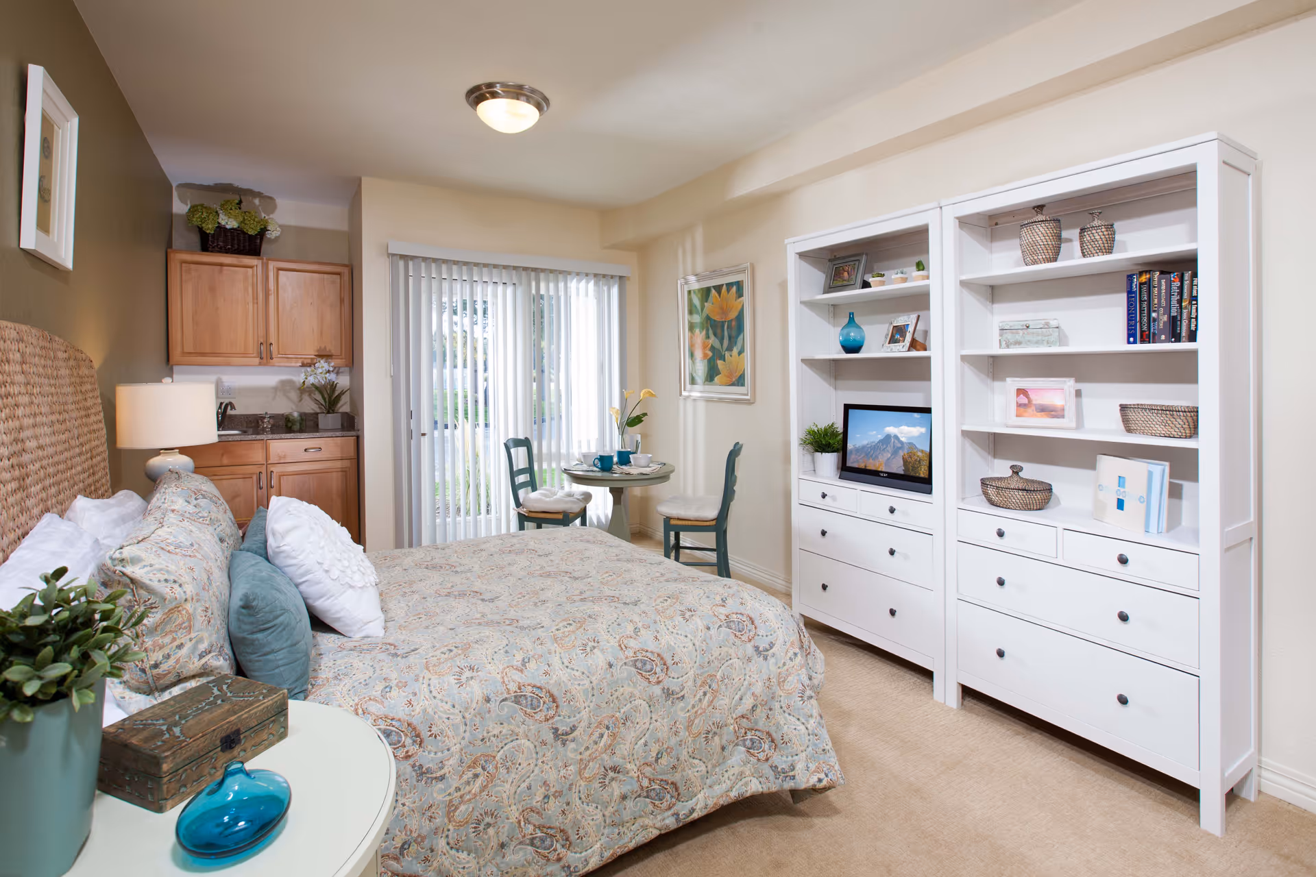 A cozy bedroom in Cottonwood Creek facility featuring a bed with patterned bedding and multiple pillows, a small round table with two chairs near a sliding glass door with vertical blinds, a white shelving unit with decorative items and a small TV, and a kitchenette area with wooden cabinets and a sink.