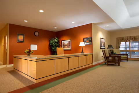 Reception area with a wooden front desk, a chair behind the desk, a lamp, framed pictures on the walls, a clock, a plant, and seating area with chairs near a window with curtains.