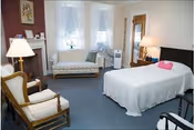 A bright furnished bedroom with a bed, armchairs, a loveseat beneath bay windows, lamps, and a small fireplace.