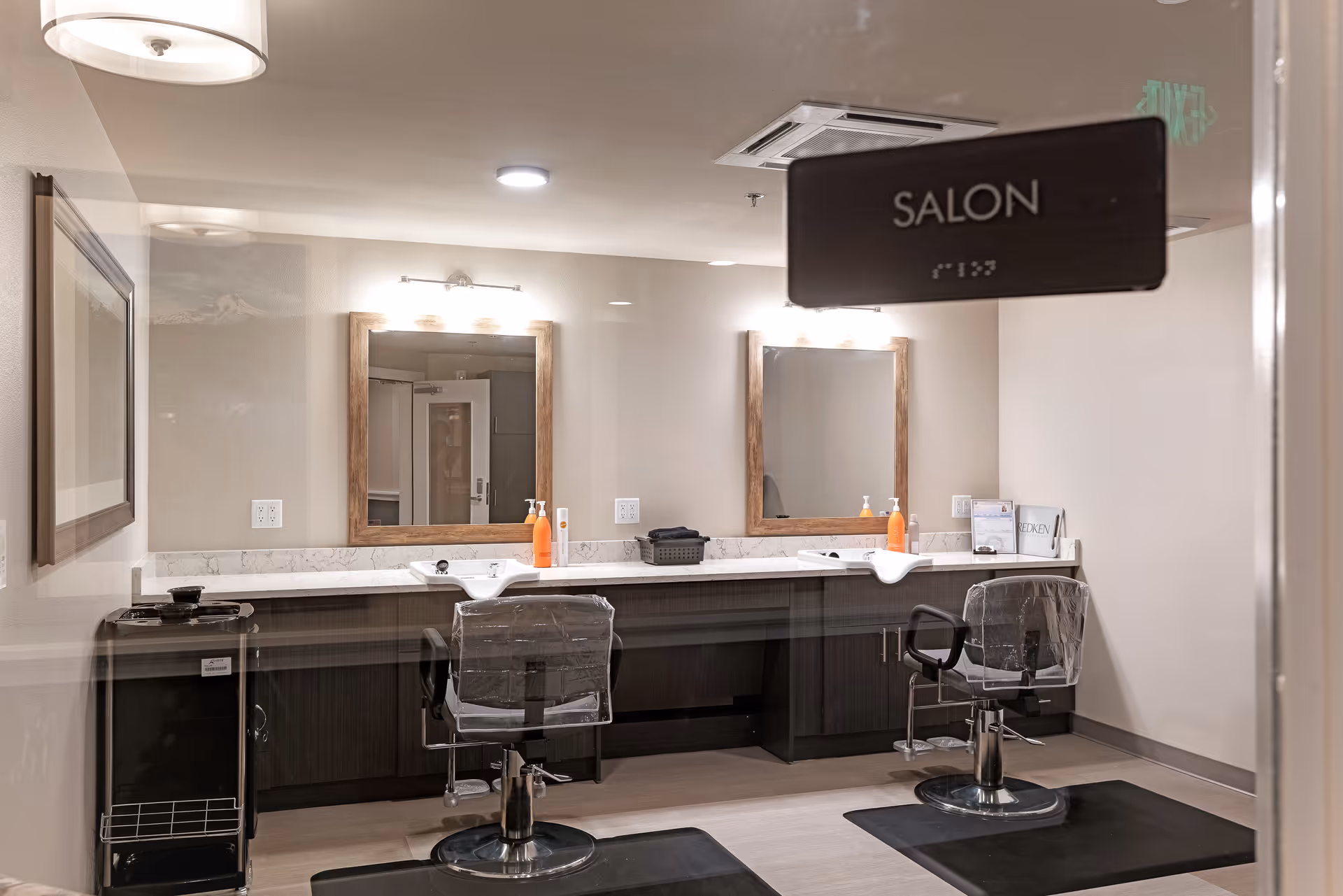 Interior view of a salon room with two styling stations, each with a mirror, a chair covered in plastic, and hair care products on the counter. The room has neutral-colored walls and flooring, with a sign on the glass door that reads 'SALON'.