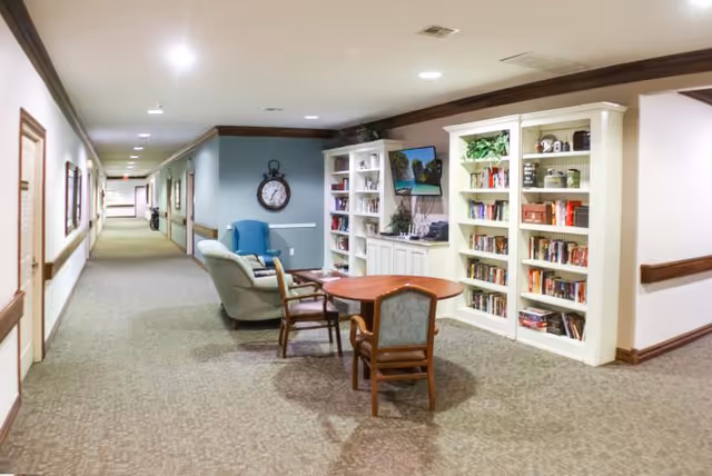 A cozy hallway area in a senior living facility featuring a round wooden table with three chairs around it, a blue armchair, a beige recliner, and two white bookshelves filled with books and decorative items. A wall-mounted TV is positioned above a cabinet between the bookshelves, and a large clock hangs on the blue accent wall. The hallway extends into the distance with doors and framed pictures along the walls.