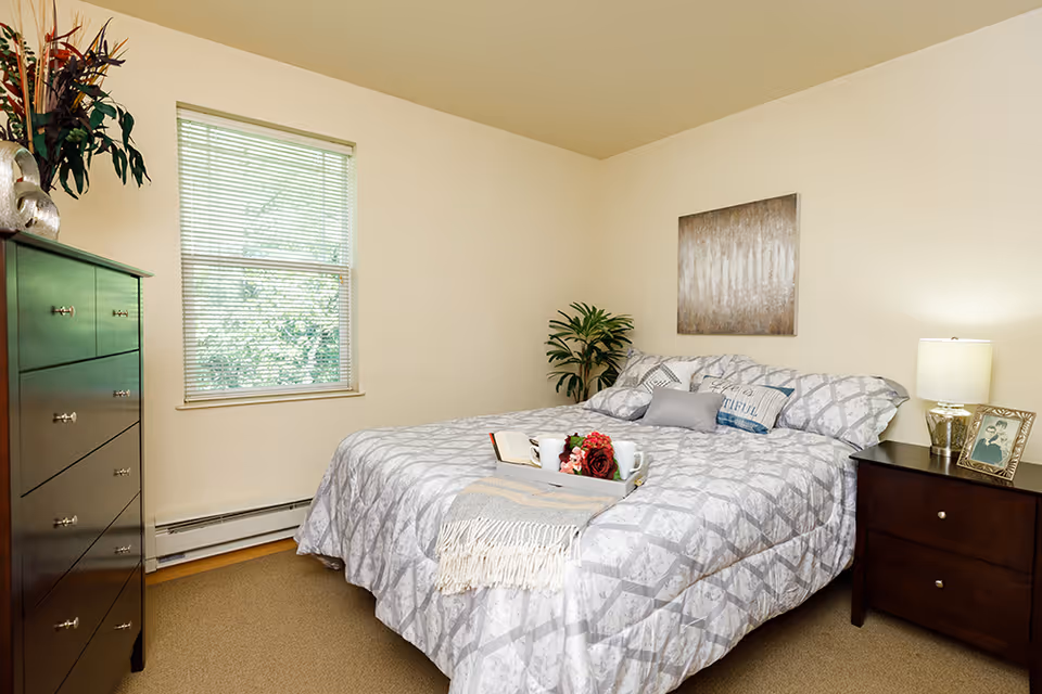 A cozy bedroom in a senior living facility with a neatly made bed featuring patterned gray and white bedding, decorative pillows, and a tray with a book, flowers, and two mugs. The room has a window with blinds, a dark wooden dresser with a plant and decorative items, a nightstand with a lamp and framed photo, and a piece of abstract wall art above the bed.