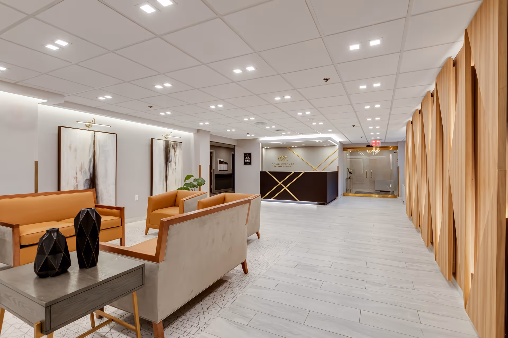A modern and spacious reception area with a seating arrangement consisting of beige and orange sofas around a small table with decorative black vases. The floor is light-colored tile, and the ceiling has recessed lighting. On the right side, there is a wooden decorative wall panel. At the far end, there is a reception desk with the Complete Care logo and name on the wall behind it.