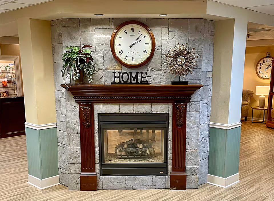 A cozy indoor fireplace with a stone surround and a dark wooden mantel. On the mantel, there is a large round wall clock with Roman numerals, a decorative plant, a metallic sunburst sculpture, and the word 'HOME' spelled out in black letters. The floor is light wood, and the walls are painted in a soft beige and green color scheme. In the background, there is a glimpse of a seating area with a clock on the wall and a lamp on a side table.