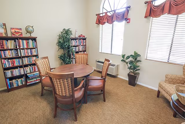 Small common room with a round wooden table and four chairs, bookshelves, plants, and windows with blinds.