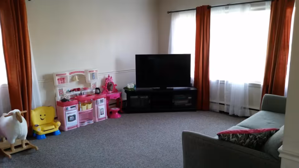 A living room with a gray sofa on the right, a large flat-screen TV on a black TV stand against the wall, and two windows with white sheer curtains and orange drapes. On the left side of the room, there is a children's play area with a pink toy kitchen set, a small pink vanity table with a mirror, a yellow smiling chair, and a white rocking horse. The floor is carpeted in gray.