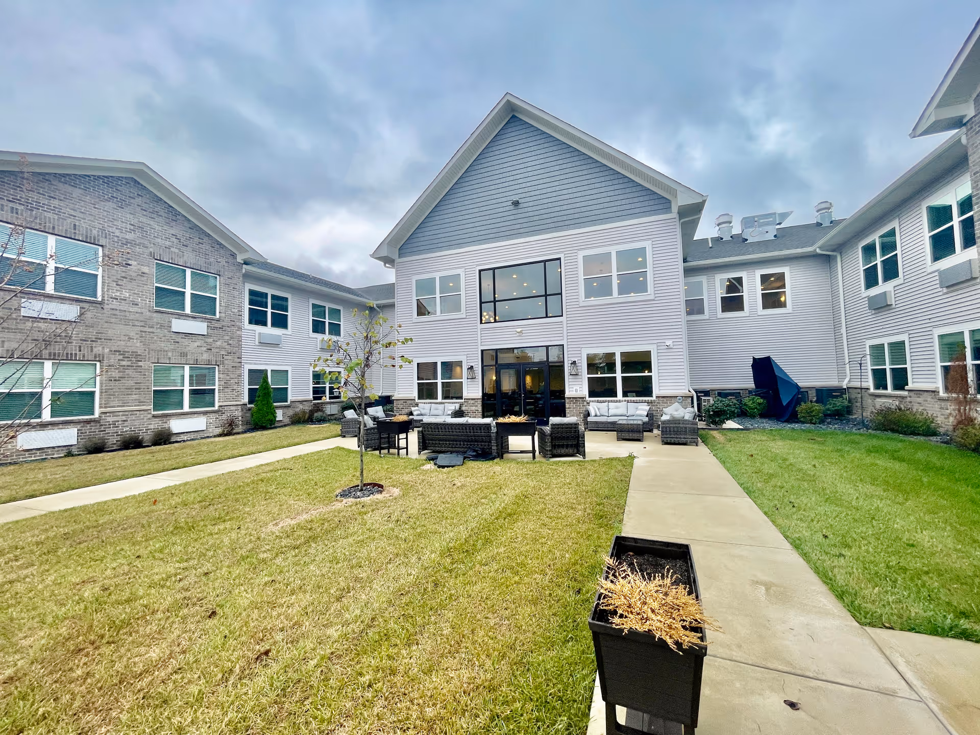 Outdoor courtyard area of Autumn Park Senior Community with a grassy lawn, paved walkways, outdoor seating with chairs and tables, and a two-story building with multiple windows surrounding the courtyard under a cloudy sky.