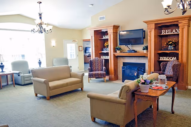A bright and cozy living room area in a senior living facility with beige sofas and armchairs arranged around a wooden coffee table with flowers and a water pitcher. The room features a fireplace with a mounted TV above it, flanked by wooden shelves displaying decorative items and words like 'RELAX' and 'WISDOM'. Large windows allow natural light to fill the space.