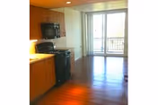 Open-plan apartment interior with a kitchen featuring wooden cabinets and black appliances opening onto a sunlit living area with sliding glass doors to a balcony.