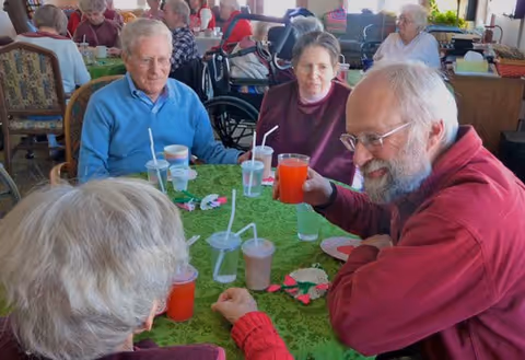 A group of elderly people sitting around a table with green tablecloth, drinking beverages through straws and engaging in conversation in a communal dining area.