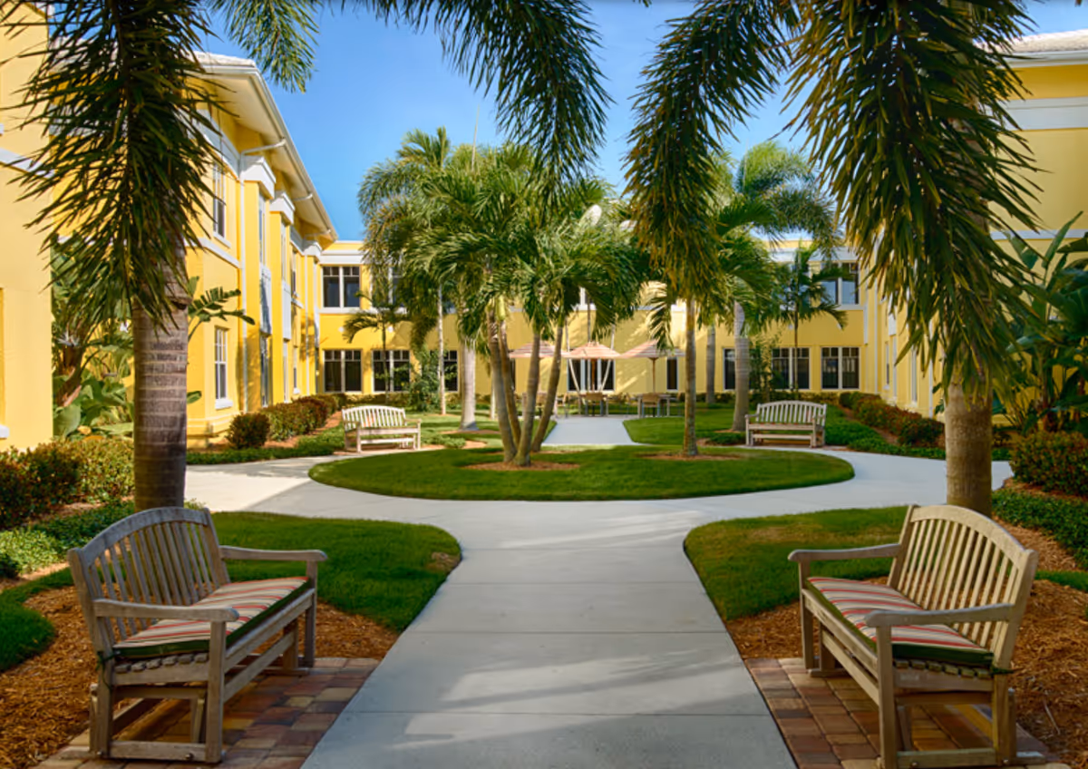 Outdoor courtyard area at The Cottage At Cypress Cove featuring a paved walkway surrounded by green grass, palm trees, and yellow buildings. There are wooden benches with striped cushions along the path and patio tables with umbrellas in the background under a clear blue sky.