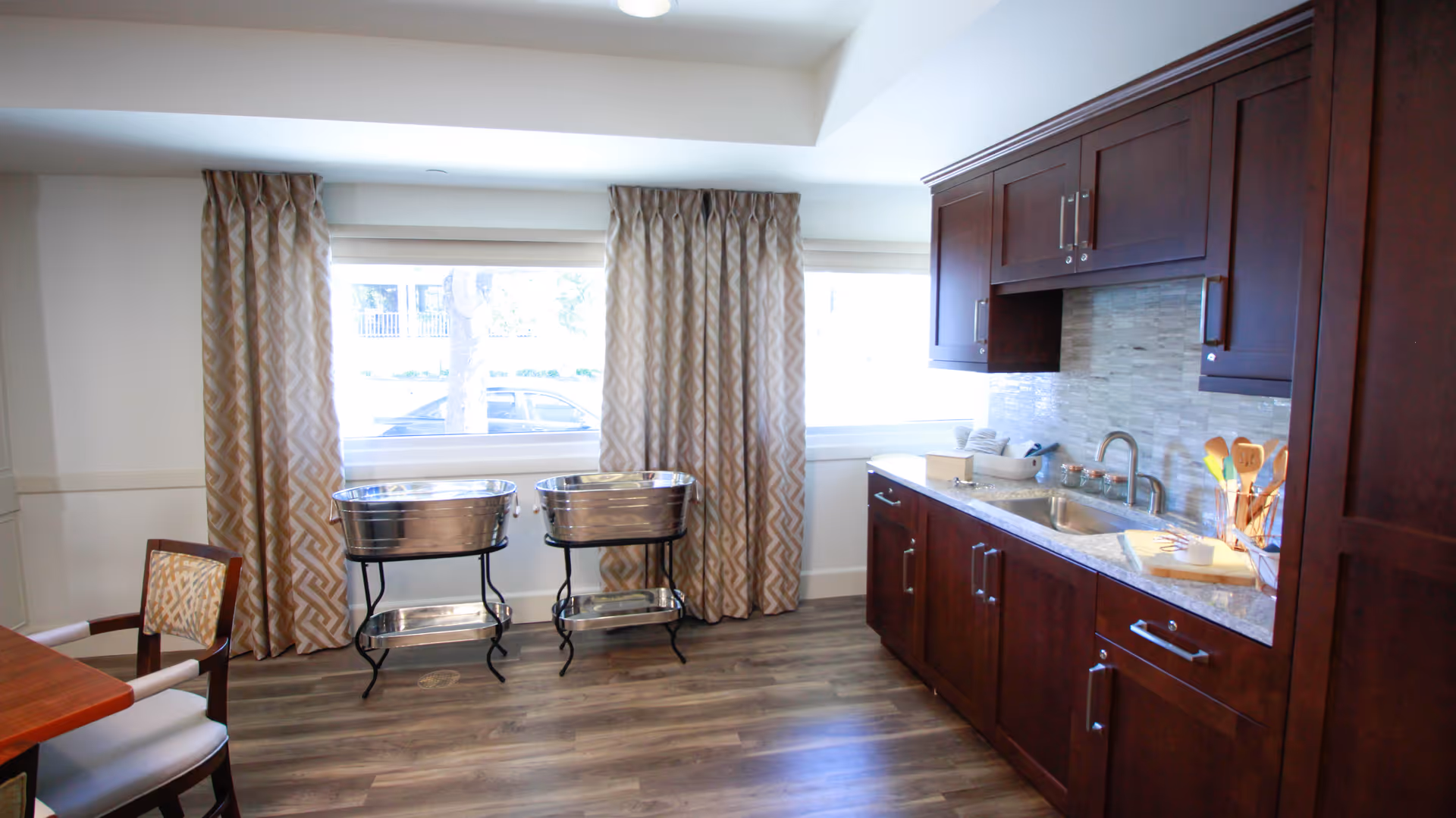 A bright room with large windows covered by patterned curtains, featuring two metal tubs on stands near the windows, a wooden dining table with chairs on the left, and a kitchen area with dark wood cabinets, a sink, and countertop on the right.
