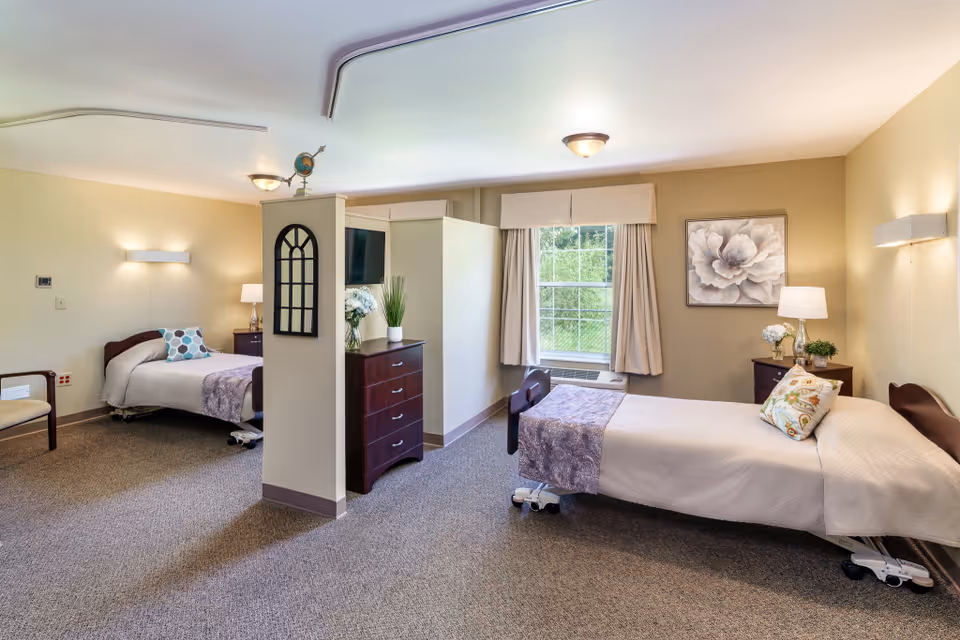 A bright and clean nursing facility room with two single beds separated by a partial wall. Each bed has a bedside table with a lamp and decorative items. The room features a large window with curtains, carpeted floor, and neutral-colored walls with a floral painting. There is a chair near one bed and a chest of drawers with plants and flowers on top.