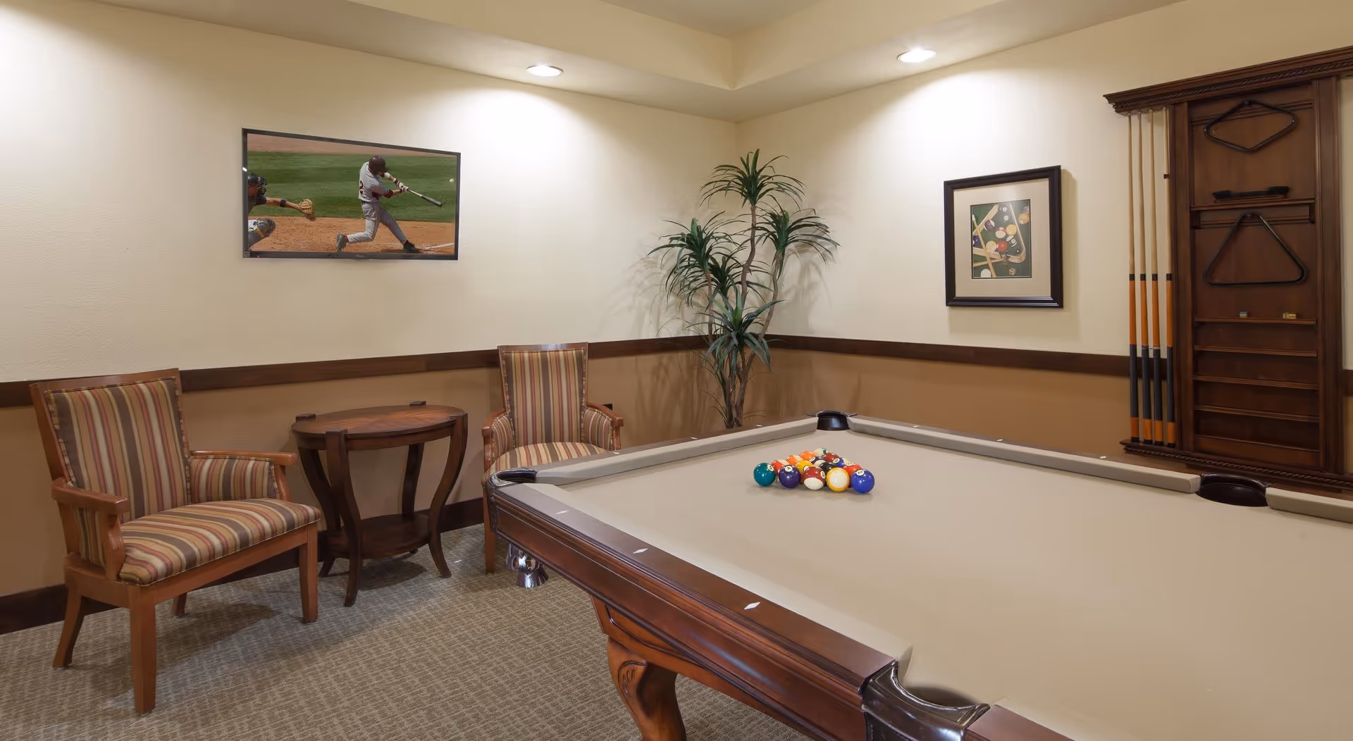 A cozy game room with a beige pool table set up for a game, two striped armchairs with wooden frames, a small round wooden table between the chairs, a tall green plant in the corner, a framed picture of billiard balls on the wall, and a flat-screen TV mounted on the wall showing a baseball game.