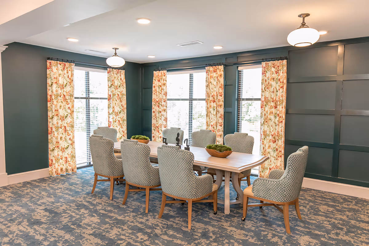 Bright dining room with a long wooden table surrounded by upholstered chairs, floral curtains, and dark green paneled walls.