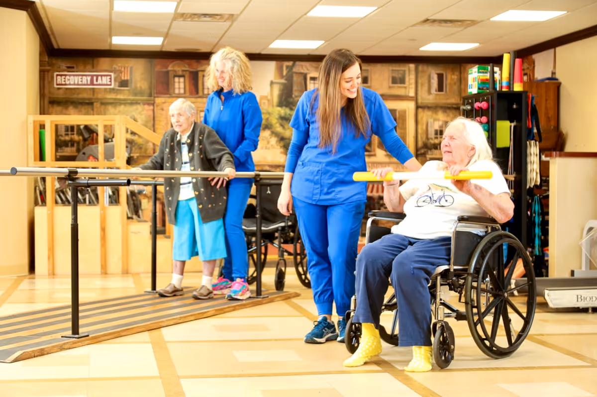 Two elderly women participating in physical therapy exercises in a rehabilitation room. One woman is using parallel bars for walking support with assistance from a caregiver in blue scrubs. The other woman is seated in a wheelchair holding a yellow exercise bar, assisted by another caregiver also in blue scrubs. The room has a mural on the wall and various therapy equipment.