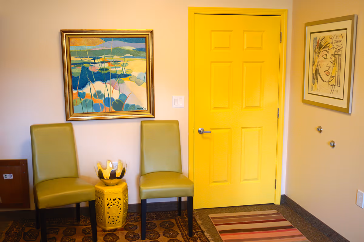 A small seating area with two green chairs and a yellow decorative side table between them. Behind the chairs is a colorful abstract painting on the wall. To the right, there is a bright yellow door and a framed artwork with a sketch of a woman's face and the words 'NO THANK YOU'. The floor has patterned rugs.