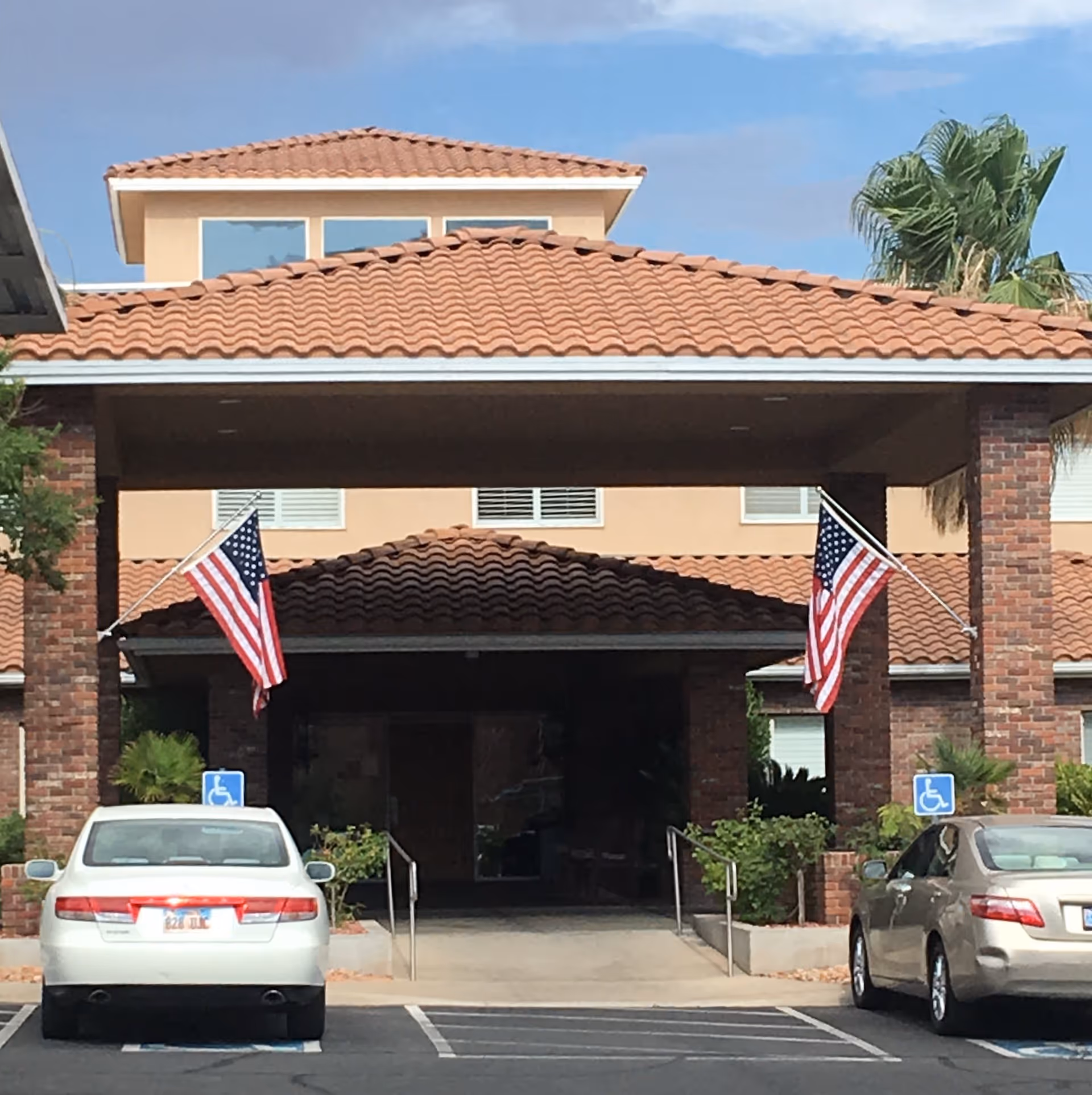 Entrance of a building with a covered driveway supported by brick pillars, two American flags mounted on the pillars, two cars parked in handicapped parking spaces in front, and a tiled roof with windows visible above.
