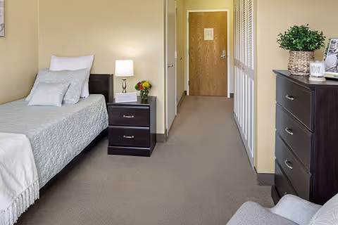 A neatly arranged bedroom in a senior living facility featuring a single bed with light blue bedding and pillows, a dark wood nightstand with a lamp and a small flower arrangement, a dark wood dresser with a plant and framed photo, beige walls, and a carpeted floor leading to a closed wooden door.